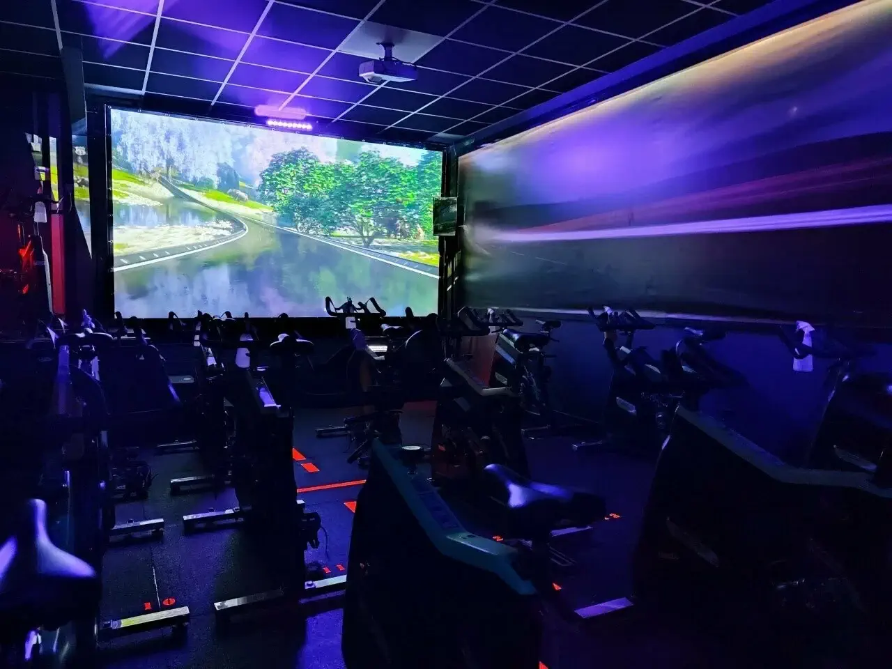 Salle de cours collectif d'indoor cycling équipée de vélos et d'un grand écran de projection immersif - Fitspro Equipement